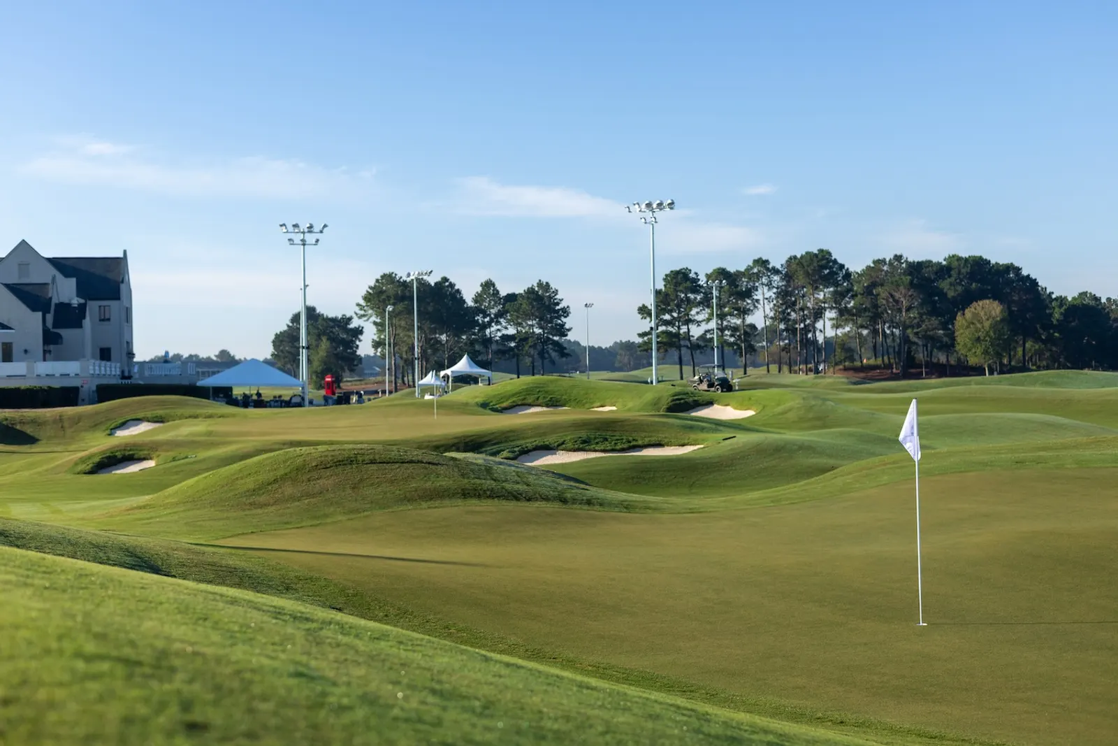 Wide view across The Piper showing mounded fairways, several bunkers, flags on two greens and tall light poles against a blue sky