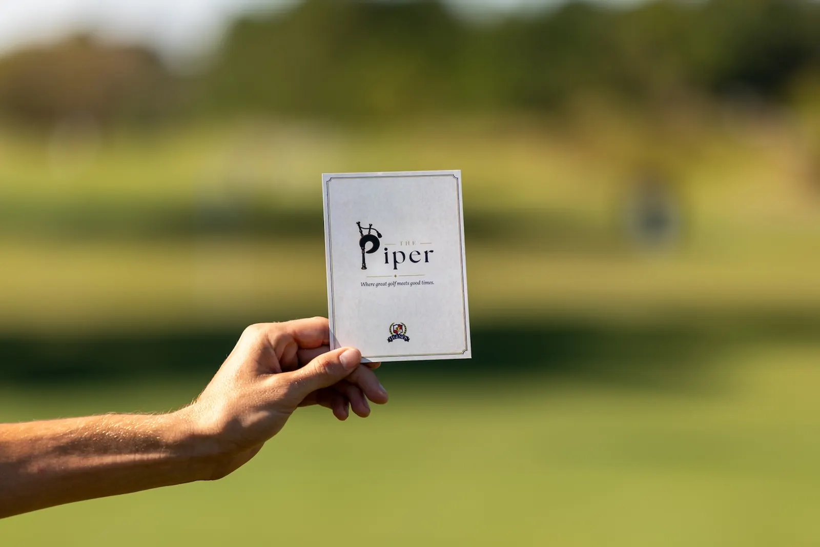 A hand holding The Piper scorecard, which reads Where great golf meets good times, with the course blurred behind