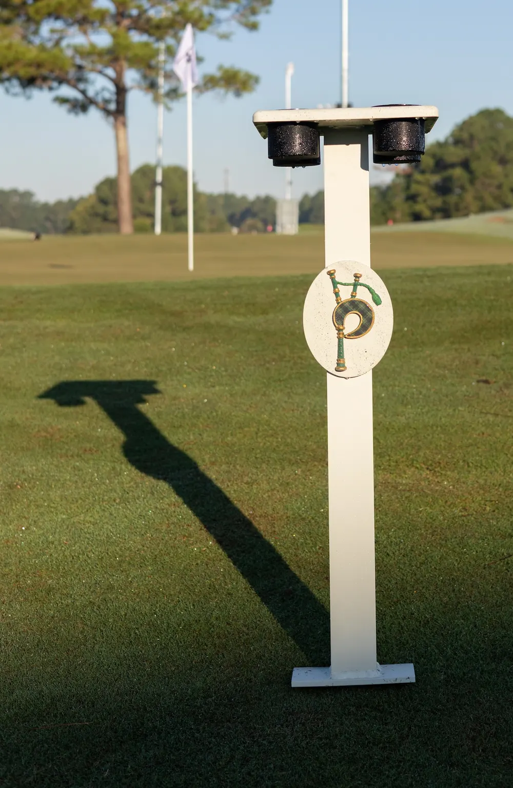 A Piper tee marker with the bagpipe logo on the tee, with the long shadow of a light pole falling across the turf