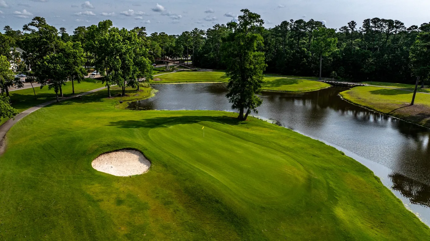 Aerial view of a River Oaks green beside a pond with a cypress tree and a wooden bridge in the distance