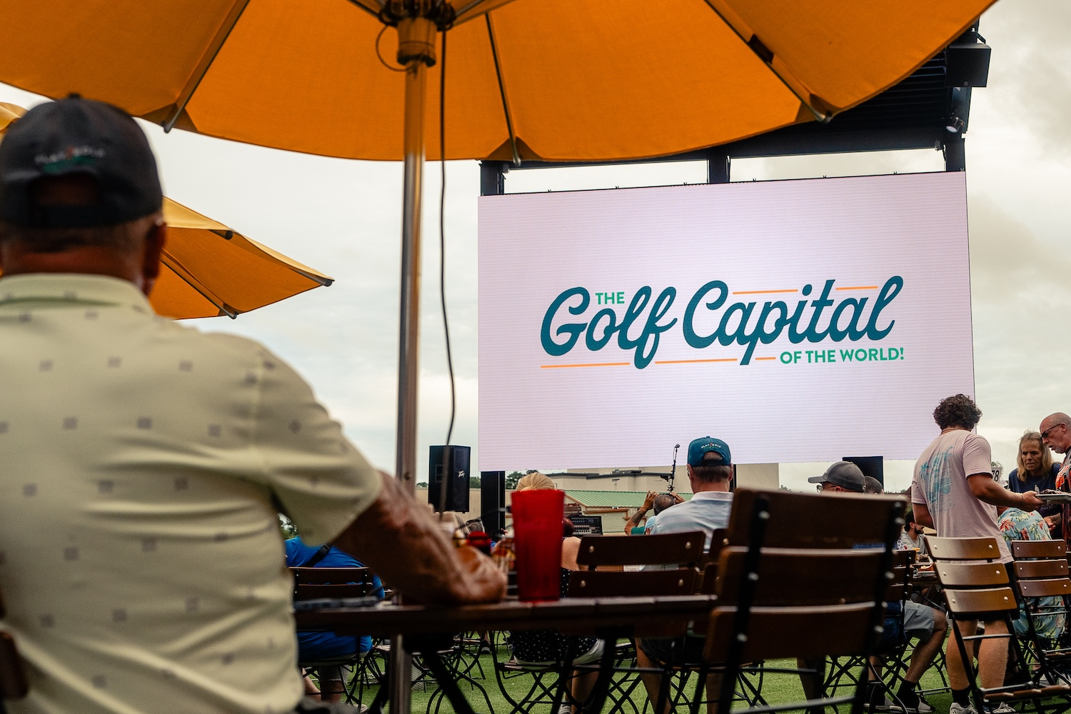 A large outdoor video screen reading The Golf Capital of the World with players seated under umbrellas