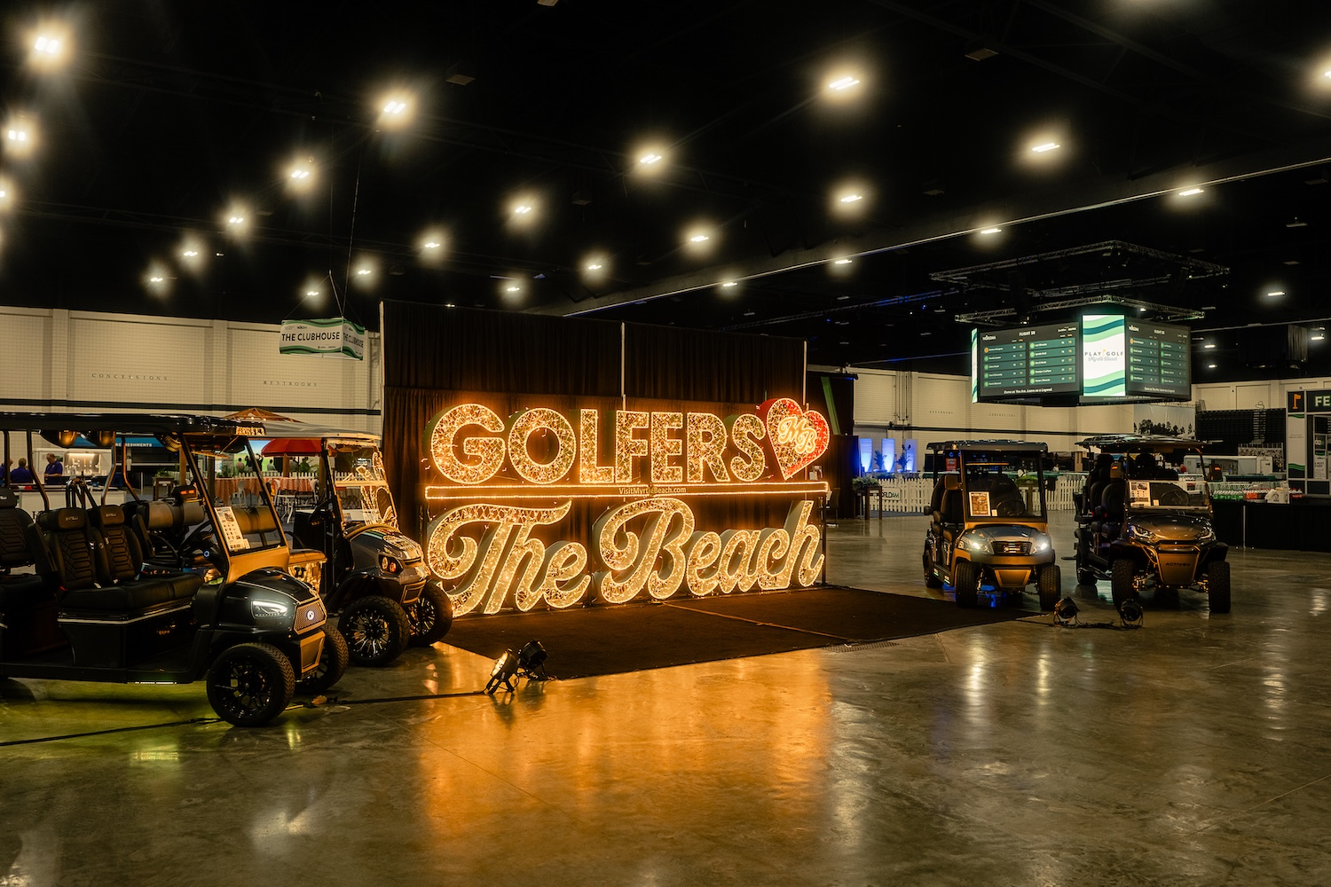 A large illuminated sign reading Golfers Love The Beach inside the convention hall with golf carts on display
