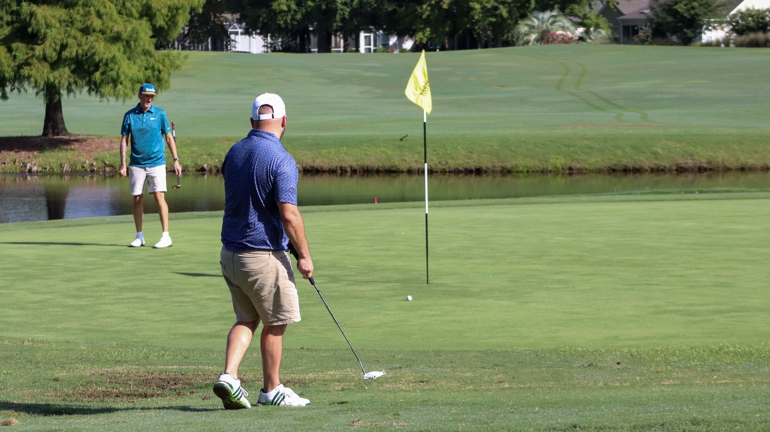 Two golfers on a green beside water, one lining up a putt in the morning sun