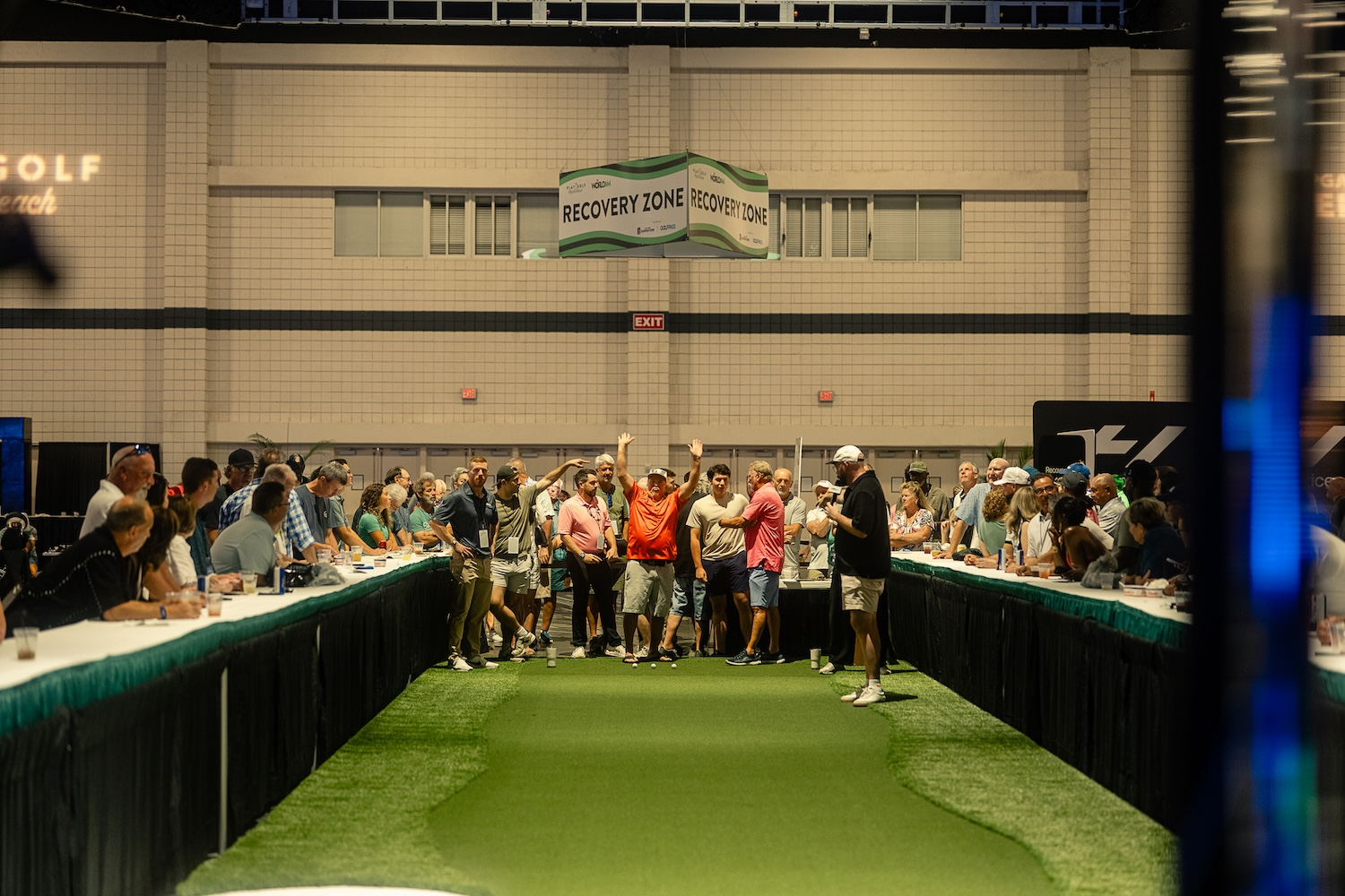 A crowd around an indoor putting lane raising their hands as a putt drops